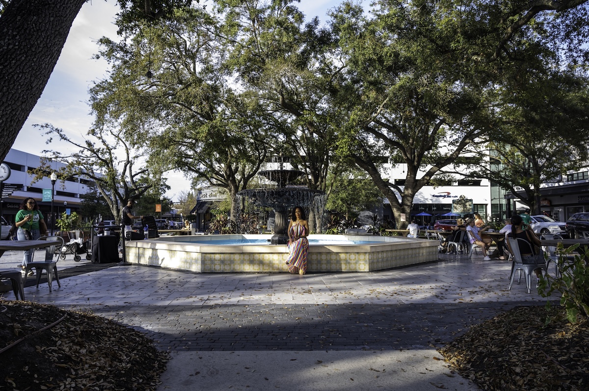 Person in a colorful striped dress standing in front of a large fountain surrounded by oak trees, outdoor seating, and shops at Hyde Park Village. This walkable shopping district offers a relaxed atmosphere for couples exploring Tampa together.