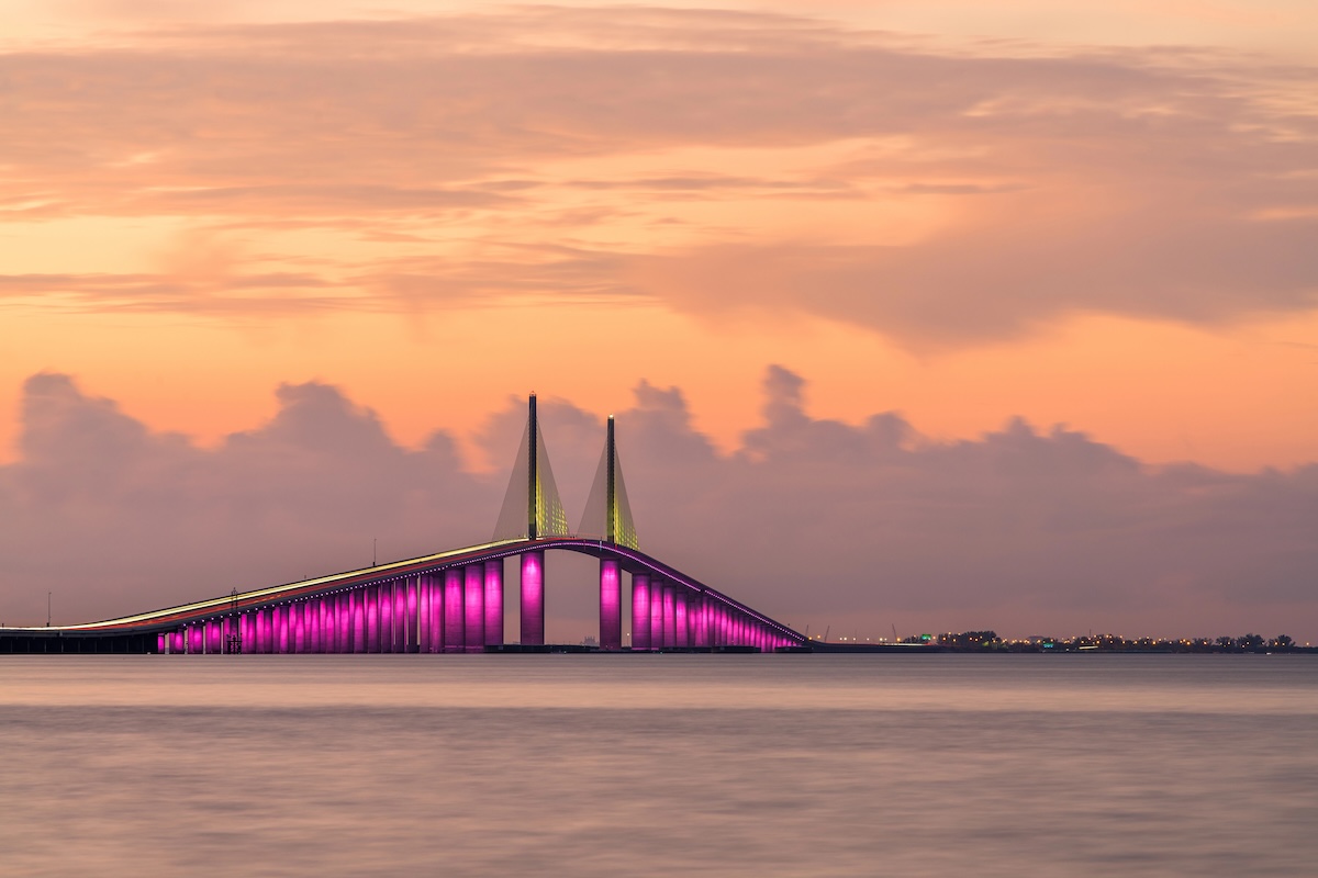 The Sunshine Skyway Bridge glows with vivid pink and yellow lights over Tampa Bay beneath a peach colored sunset sky. Calm water fills the foreground as the illuminated bridge stretches toward the horizon.