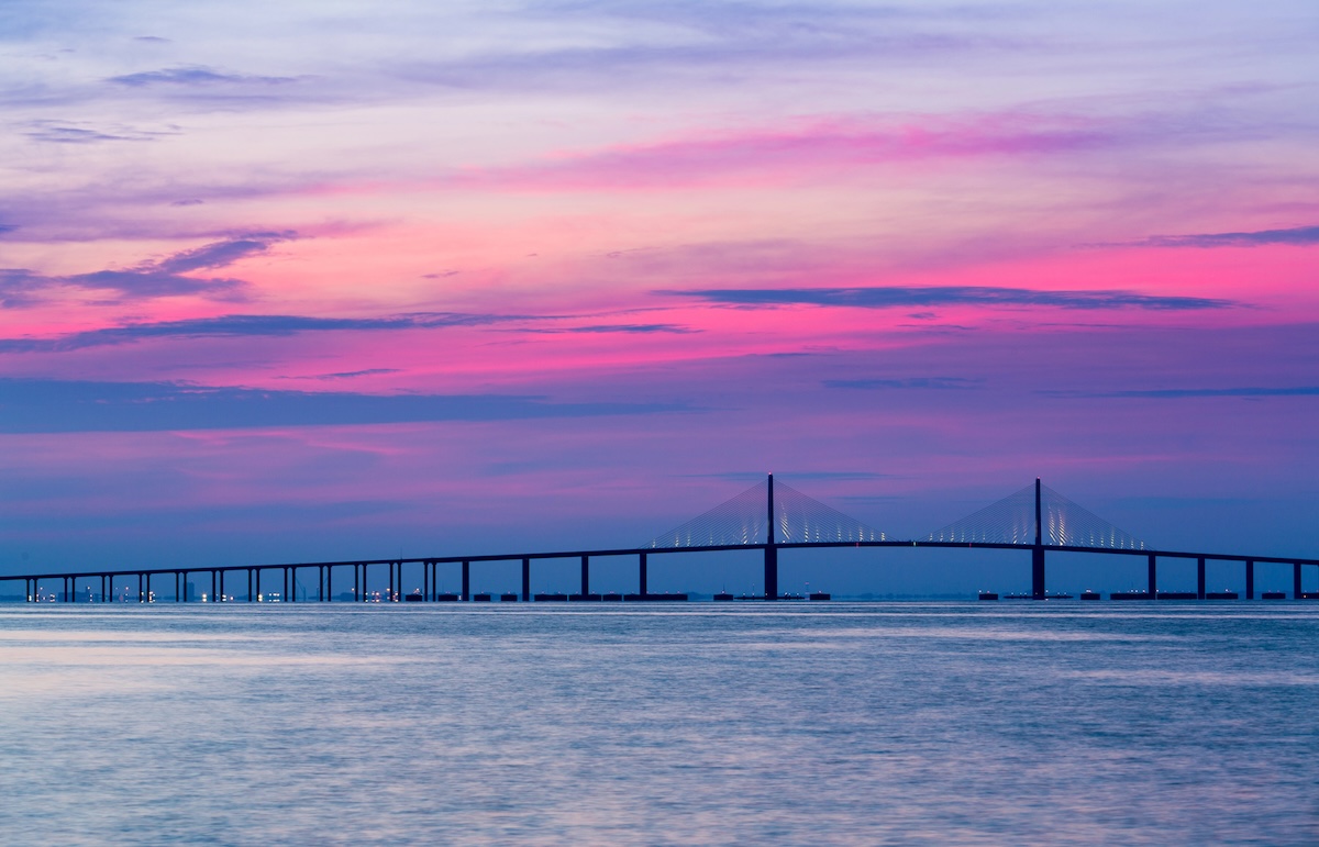 The Sunshine Skyway Bridge stretches across Tampa Bay beneath a dramatic pink purple and blue twilight sky. The bridge and colorful sunset create a scenic view from the water.