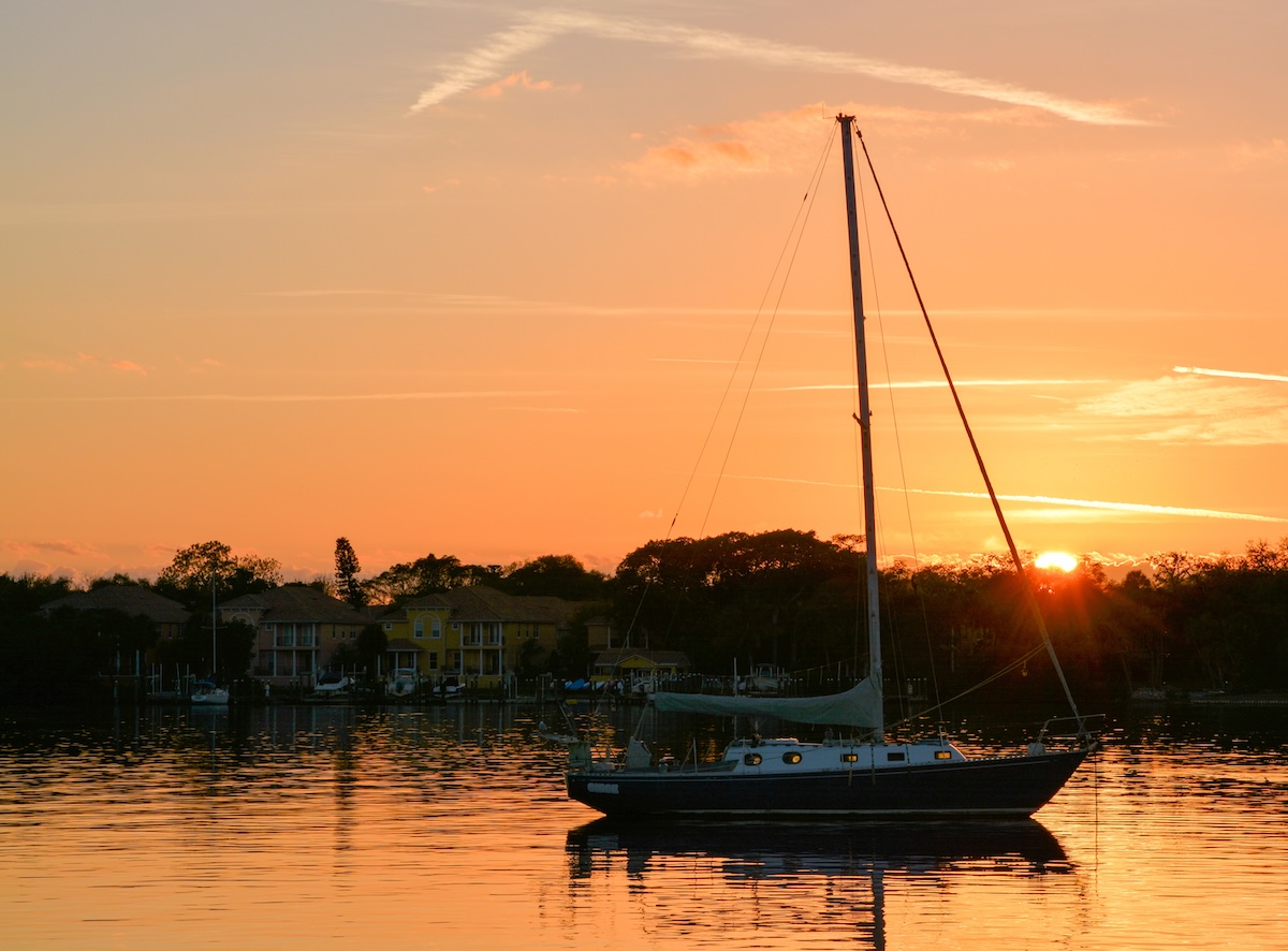A sailboat rests on calm water as the setting sun glows orange behind waterfront homes and trees. Warm sunset colors reflect across the water around the silhouetted boat.