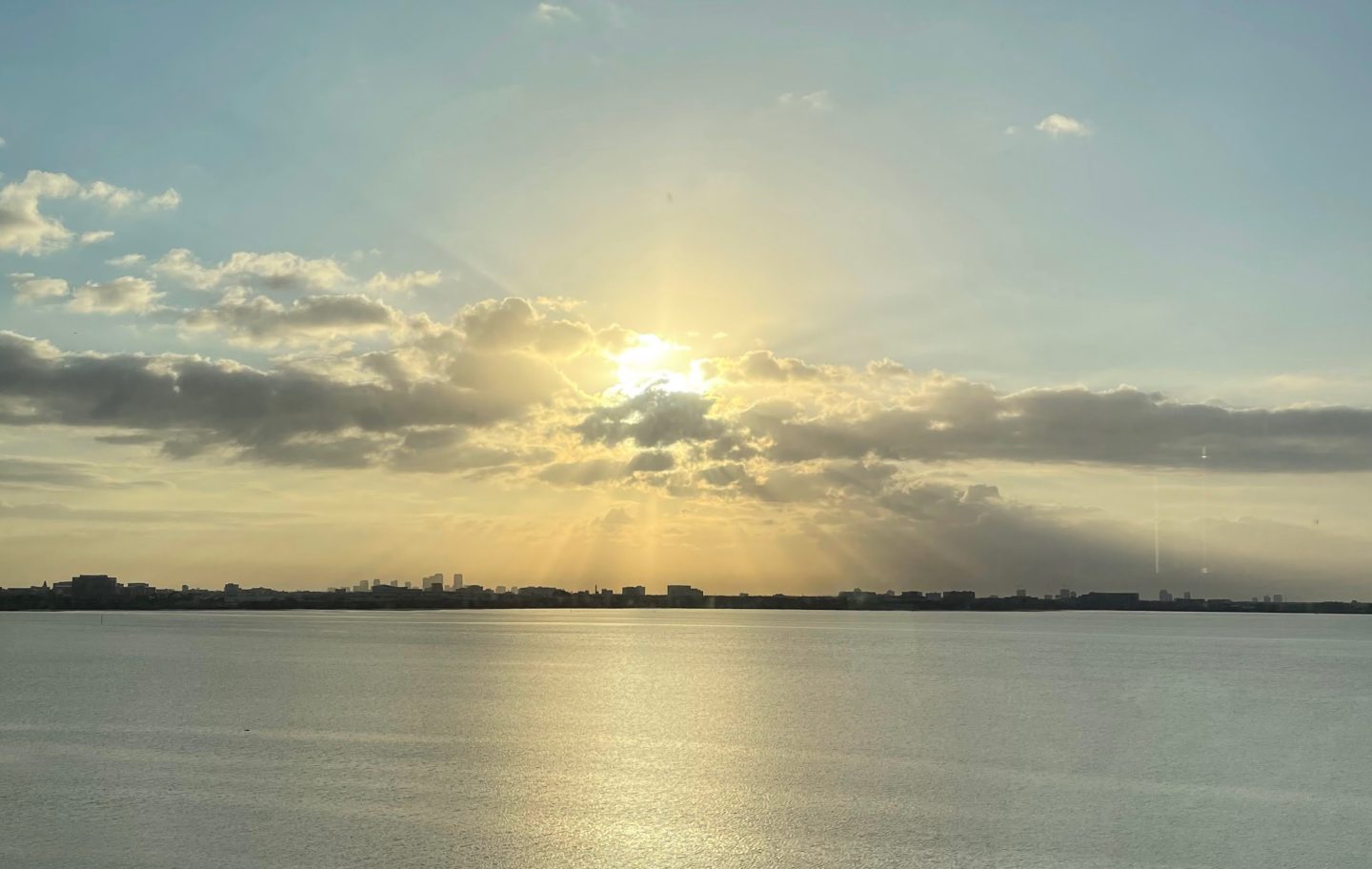 Golden sunlight breaks through layers of clouds over Tampa Bay and reflects across the calm water. The distant Tampa skyline is silhouetted along the horizon as the sun begins to set.