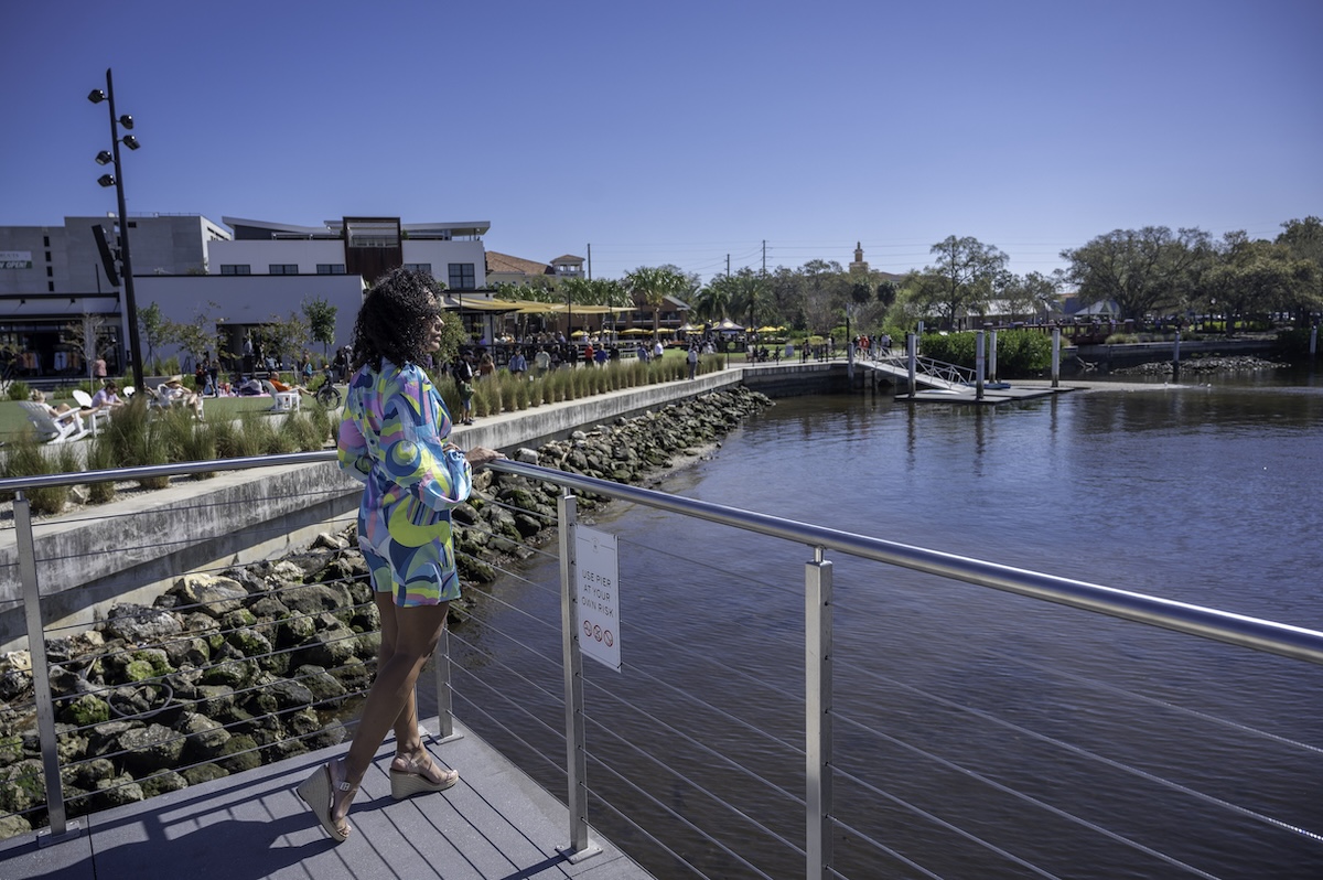 Person standing on a waterfront boardwalk overlooking the Hillsborough River with restaurants, green space, and a marina in the background. The scenic Riverwalk area is a popular choice when deciding where to stay in Tampa for couples.